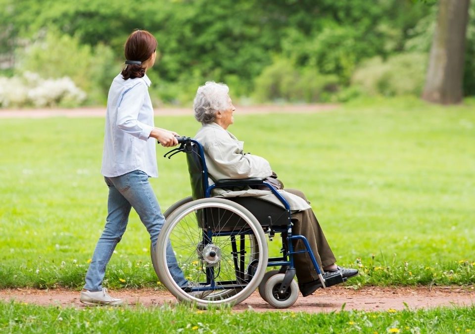 Une personne agée en fauteuil poussée par une aide soignante dans un parc.